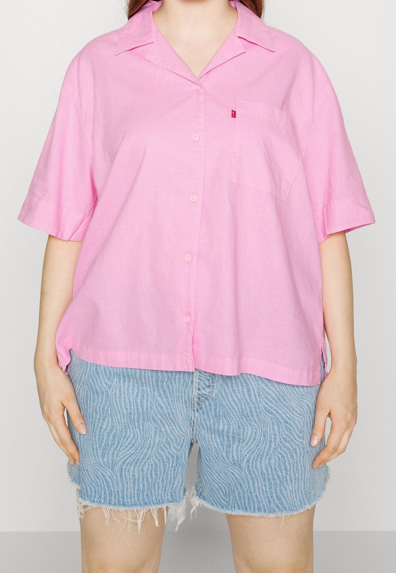 Personne portant une chemise rose ample à manches courtes avec des boutons et un short en jean bleu clair à motifs avec des bords effilochés.