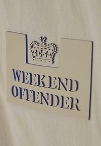 Beige stofbaggrund med et cremefarvet mærke, der har blå tekst "WEEK END OFFENDER" og et kronegrafik ovenfor.