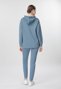 Γυναίκα με καστανά μαλλιά σε κότσο, φορώντας μπλε φούτερ με κουκούλα και αντίστοιχο παντελόνι, όρθια και με την πλάτη προς τα εμπρός, σε απλό φωτεινό φόντο.