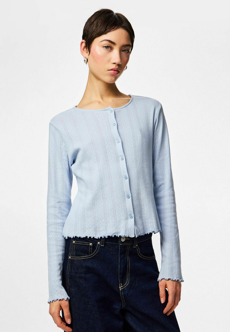 Νεαρή γυναίκα που φοράει ανοιχτό μπλε ζακέτα με κουμπιά και ριμπ υφή, συνδυασμένη με σκουρό μπλε τζιν, στέκεται μπροστά σε απλό λευκό φόντο.