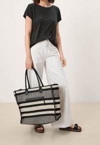 Sac fourre-tout rayé en matériau noir et beige, de forme rectangulaire, avec deux poignées, présentant une poche à l'avant. Porté avec un pantalon large blanc.