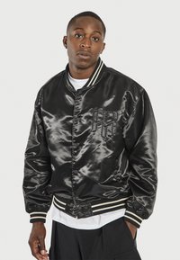 Chaqueta bomber de satén negra con puños y cinturilla de canalé a rayas, que cuenta con un cierre de botones y un logotipo bordado en el pecho.