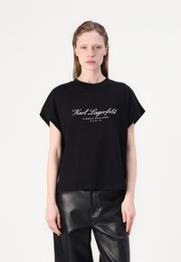Juoda trumpomis rankovėmis marškinėliai su baltu užrašu "Karl Lagerfeld 21 RUE ST.-GUILLAUME PARIS". Minkšta medžiaga, laisvas kirpimas ir apvalus iškirpimas.