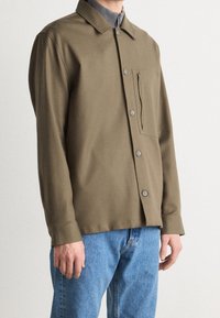 Chaqueta de botón verde oliva hecha de una tela suave, con un bolsillo en el pecho, y combinada con jeans de color azul claro.