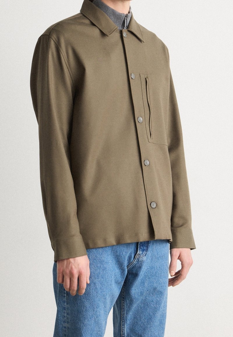 Chaqueta de botón verde oliva hecha de una tela suave, con un bolsillo en el pecho, y combinada con jeans de color azul claro.