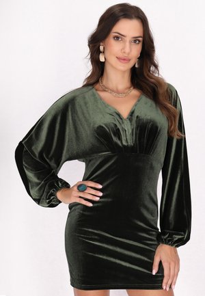 Woman with long dark hair wears a dark green velvet dress with long puffed sleeves and gold earrings and necklace, posing with one hand on hip.
