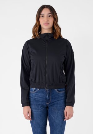 Giacca zip-up nera con collo alto, vita elastica e due tasche laterali. Indossata sopra jeans in denim blu. Tessuto liscio e leggero.