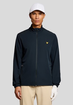 Uomo che indossa una giacca nera con zip e logo giallo, pantaloni beige, guanto da golf bianco alla mano sinistra e cappellino da baseball bianco traforato.