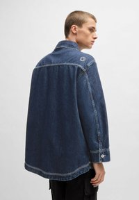 Donkerblauwe denimjas met een losse pasvorm, spitse kraag en knoopmanchetten. Heeft een klein logo detail op de schouder.