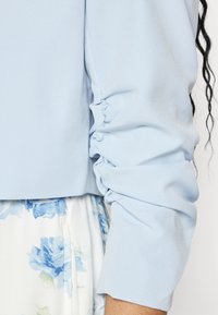 Blouse bleu clair avec manches froncées. Tissu lisse avec une texture légèrement brillante. En dessous, une jupe à motif floral en bleu et blanc.