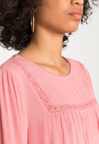 Blusa rosa texturizada com gola arredondada e detalhes em renda ao longo da blusa, com tecido solto e fluido e ombros caídos.