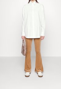 Vitskjorta med långa ärmar, tanfärgade korrugerade utsvängda byxor och en liten väska med ormskinnsmönster. Vita sneakers med bruna detaljer.