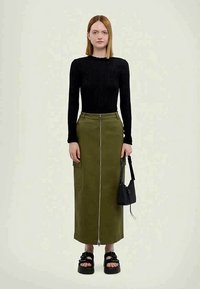 Femme portant un haut noir à manches longues, une jupe taille haute olive verte avec fermeture éclair à l'avant, des sandales à plateforme noires, et tenant un petit sac à main noir.