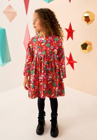 Robe rouge avec des motifs floraux et de fêtes, manches longues, taille froncée, associée à des bottes noires montantes jusqu'aux genoux et des leggings noirs.