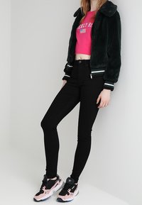 Chaqueta bomber de piel sintética verde, sudadera corta rosa con texto, jeans ajustados negros y zapatillas negras/rosas con suela gruesa.