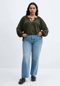 Femeie stand cu mâinile în buzunare, îmbrăcată cu o bluză verde măslin, blugi albaștri deschiși și pantofi slip-on negri cu ținte.