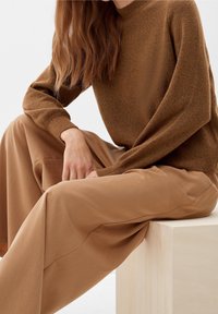 Pull marron texturé à manches longues, associé à un pantalon kaki à jambes larges. Le mannequin est assis sur un bloc en bois clair.