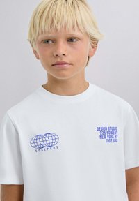 Weißes T-Shirt aus Baumwolle mit blauen Grafikprints: ein Globus-Logo auf der linken Brust und Text auf der rechten Seite. Kurze Ärmel.