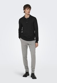 Schwarzes Langarm-Poloshirt mit Knopfleiste, kombiniert mit hellgrauen, schmal zulaufenden Hosen und schwarzen Stiefeletten. Glatte Stoffstruktur.