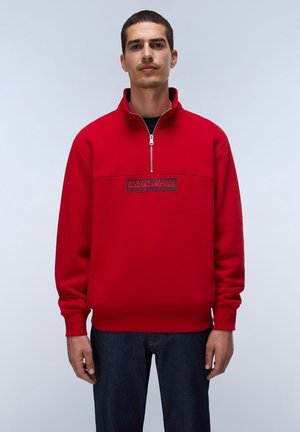 Mann steht und trägt ein rotes Quarter-Zip-Sweatshirt mit einem Logoaufnäher auf der Brust und dunkelblaue Jeans vor einem schlichten, hellen Hintergrund.