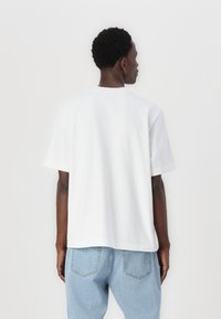 Tricou alb cu maneci scurte din bumbac, cu un croi lejer și un gât rotund. Vederea din spate arată un design minimalist și o textură netedă.