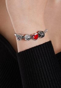 Zilveren charm armband met hartvormige bedels, een rode emaille hart, een bloemmotief en een fonkelende steen, gedragen om een pols.