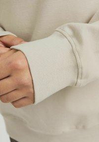 Beige pamut pulóver bordás mandzsettákkal. Simaságának köszönhetően minimalista dizájn, látható varratok az éleken. Közelkép a csukló területéről.