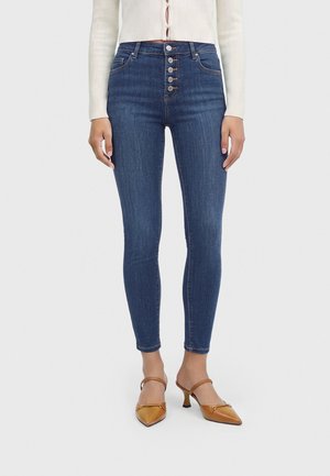 Femme portant un jean skinny bleu foncé taille haute avec cinq boutons sur le devant, associé à un pull court blanc et des chaussures marron à talons.