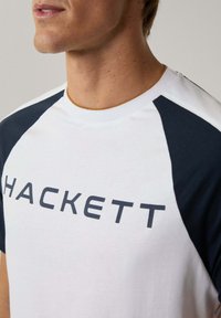 Biela tričko s námorníckymi modrými raglánovými rukávmi, okrúhlym výstrihom, s nápisom značky "HACKETT" tučným tmavým písmom naprieč hrudníkom.