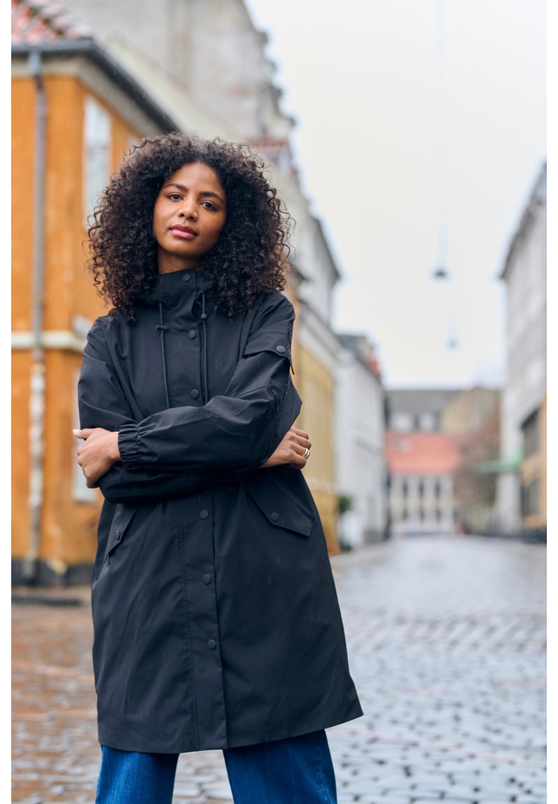 Parka noire imperméable avec un col haut, une capuche à cordon de serrage et des poignets élastiques. Comprend des poches avant et un design épuré et minimaliste.
