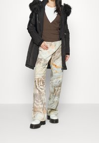 Abrigo acolchado negro con capucha de pelo sintético, blusa envolvente marrón y pantalones de pierna ancha con un patrón marmoleado en tonos beige y azul, acompañados de botas blancas gruesas.
