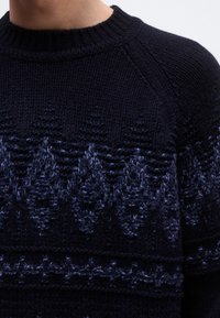 Tengerészkék kötött pulóver világosabb árnyalatú geometriai mintával, bordázott körkivágással és raglán ujjakkal. Texturált fonal részletek.