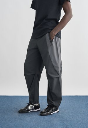 Asmuo vilki tamsiai pilkas kelnes su užtrauktukais šoninėse kišenėse ir juodus sportinius batelius su baltomis pado dalimis, stovintis ant mėlyno kilimo.