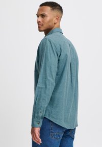 Chemise à manches longues de couleur sarcelle en tissu texturé, avec poignet boutonné et détail de pli dans le dos, portée avec un jean bleu.