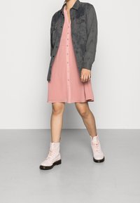 Grijze button-up jasje over een roze button-front jurk, gecombineerd met lichtroze veterschoenen tot de enkel. Glad materiaal en gestructureerd ontwerp.