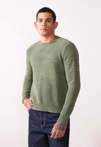 Green knit sweater with a textured pattern, crew neckline, and ribbed cuffs. Paired with dark blue denim jeans.