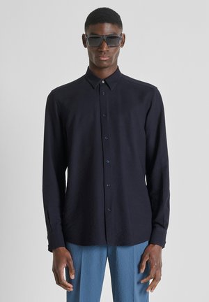 Man draagt een zwarte overhemd, blauwe gestructureerde broek en donkere zonnebril, staand tegen een effen lichtgrijze achtergrond.