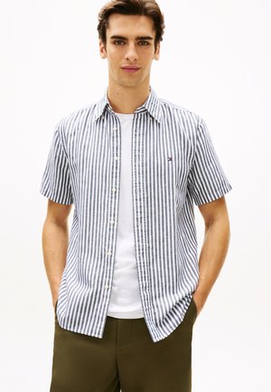 Νεαρός άνδρας με τα χέρια στις τσέπες, φορώντας πουκάμισο με κοντά μανίκια σε κάθετες ριγέ μπλε και λευκό, πάνω από λευκό T-shirt και χακί παντελόνι.