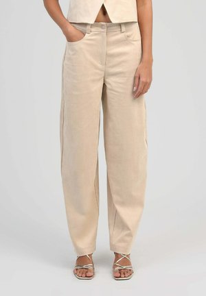 Pantalon beige taille haute à jambes larges porté avec des sandales argentées à brides, une main dans la poche, top court laissant voir le ventre.