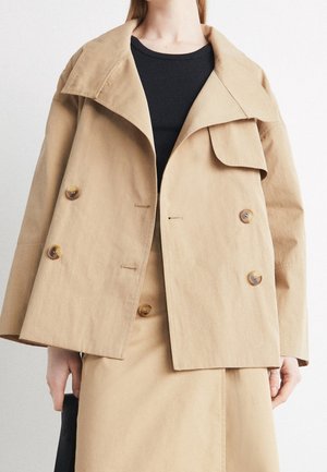 Femme portant une veste beige double boutonnage sur un haut noir et une jupe beige assortie, tenant un sac noir dans la main gauche.