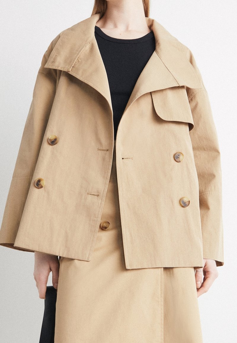 Femme portant une veste beige double boutonnage sur un haut noir et une jupe beige assortie, tenant un sac noir dans la main gauche.