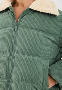 Zipped green corduroy jacket with cream fleece collar worn, showing textured fabric and hand resting on hip.
