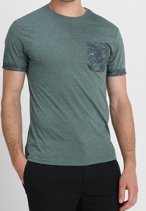 Mann trägt ein grünes T-Shirt mit kurzen Ärmeln, einer dunklen Tasche und Ärmelbündchen mit Blattmuster, kombiniert mit einer schwarzen Hose.