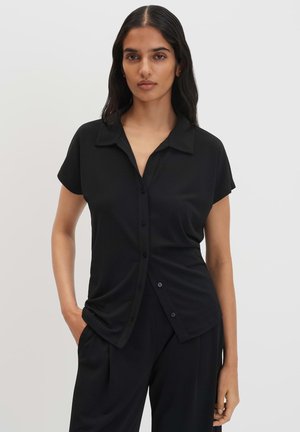 Vrouw met lang donker haar, gekleed in een zwart overhemd met korte mouwen en zwarte broek, staand met één hand in de zak.