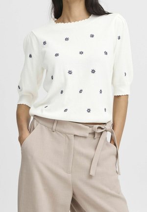 Femme portant un haut à manches courtes en maille blanche avec des étoiles brodées noires, rentré dans un pantalon beige taille haute avec une ceinture en tissu nouée en nœud.