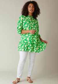 Tunic floreale verde con motivi di margherite bianche, colletto, maniche lunghe e orlo arricciato, abbinata a pantaloni slim-fit bianchi e sandali.