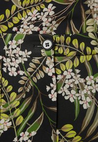 Tissu à motifs floraux noirs, avec des fleurs blanches texturées, des accents roses et des feuilles vertes. Comprend un bouton noir en détail.
