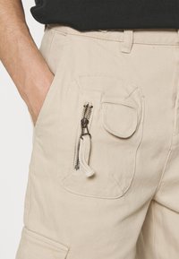 Bežové cargo kalhoty mají boční kapsu na zip a dekorativní třásně. Materiál má texturovaný povrch s viditelnými detaily šití.
