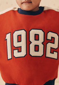 Pull rouge arborant le chiffre « 1982 » en crème avec des accents bleu marine. Texture douce et col côtelé ; un design décontracté et sportif.