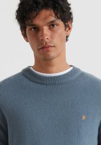 Niebieski sweter z dzianiny z okrągłym dekoltem, z małym pomarańczowym logo na klatce piersiowej. Gładka tekstura i lekka konstrukcja.